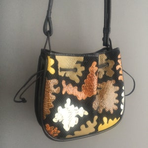 May include: A black leather bucket bag with a drawstring closure. The bag is decorated with colorful, geometric shapes in shades of brown, gold, and white.