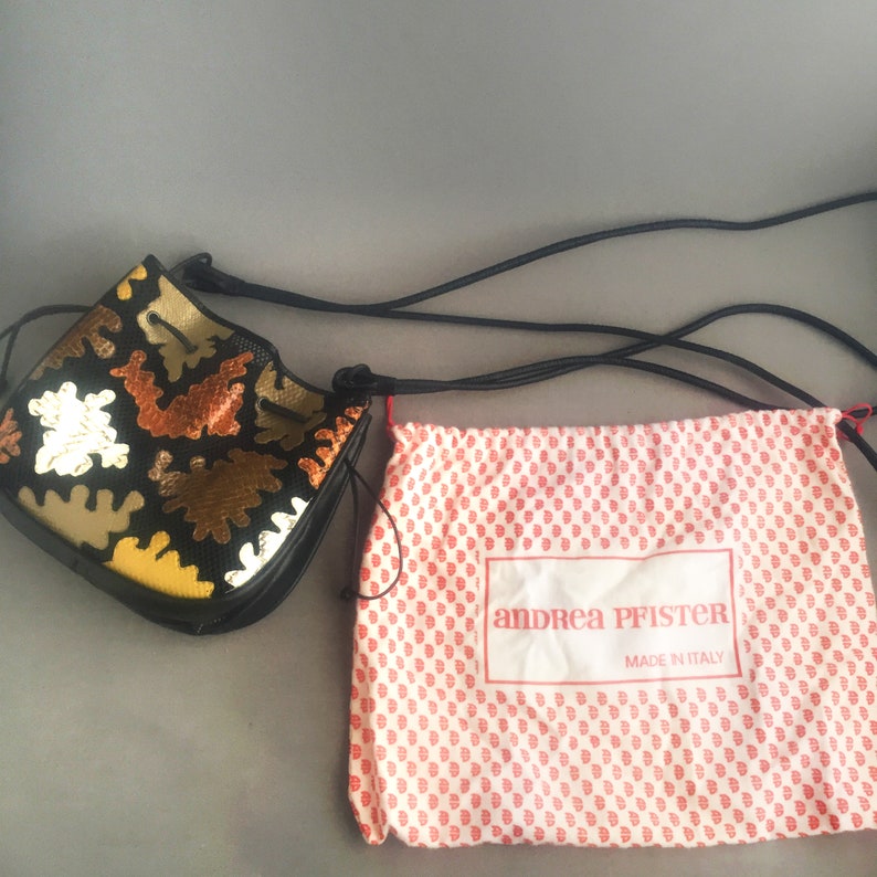 May include: A black bucket bag with a colorful geometric pattern and a drawstring closure. The bag comes with a dust bag that has the text "andrea pfister made in italy" printed on it.