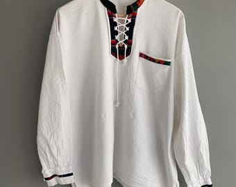 May include: White long-sleeved shirt with a lace-up neckline and a colorful patterned trim. The shirt has a single pocket on the left side.