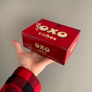 May include: A vintage red OXO cubes box with white lettering. The box reads "OXO cubes" and "For cooking and drinking". The box is held in a hand and has a distressed appearance.