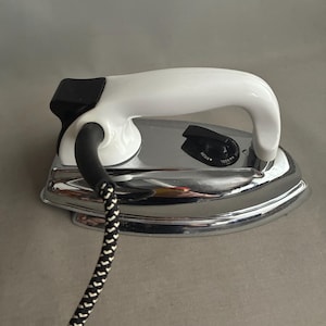 Puede incluir: Una plancha eléctrica vintage con mango blanco y base cromada. Cuenta con un dial de temperatura negro y un cable con diseño en blanco y negro. Un electrodoméstico clásico.
