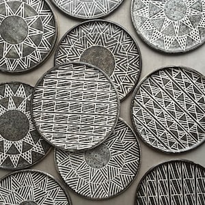 May include: A collection of dark gray ceramic plates with white geometric patterns. The plates feature various designs, including sunbursts, zigzags, and linear motifs. The plates are arranged on a gray surface, showcasing their decorative appeal.