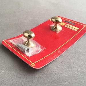 May include: Two brass-colored cabinet knobs on a red and yellow rectangular card. One knob is packaged with a screw in a clear plastic bag. The card has the text "Easy to fit" and "All things included".