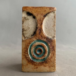 Puede incluir: Un jarrón rectangular de cerámica con una superficie marrón terrosa texturizada. Presenta dos hendiduras blancas semicirculares en la parte superior y un diseño de círculos concéntricos turquesa en el centro. El jarrón tiene una estética minimalista y abstracta.