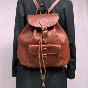 Op de afbeelding: Een bruine leren rugzak met een voorvak en een trekkoord. De rugzak heeft een leren band en een metalen gesp.