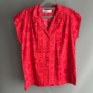 Peut inclure: Une chemise rouge à manches courtes avec un motif répétitif de motifs floraux, géométriques et d'ananas. La chemise a un col classique et une fermeture à bouton unique. L'étiquette indique "Sea Shells".