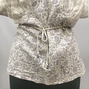 May include: A white blouse with a purple floral pattern and a tie at the waist. The tie has small gold beads at the ends.