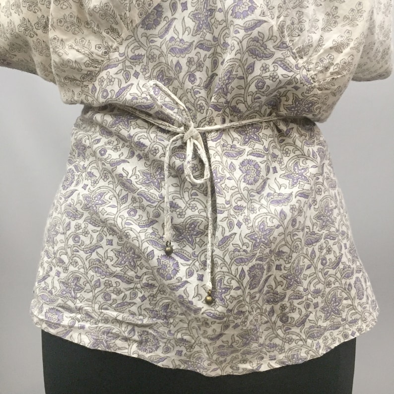 May include: A white blouse with a purple floral pattern and a tie at the waist. The tie has small gold beads at the ends.