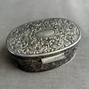 May include: Oval silver-toned box with intricate floral and scrollwork patterns. The box has a hinged lid and a small, oval design on the top. The metal box is likely made of silver or a silver alloy.