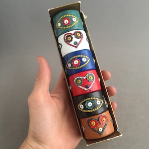 May include: Set of six colourful napkin rings with embroidered designs. Each ring features a different colour and pattern, including red, blue, green, white, and black. The rings are made of wood and have a vintage look.