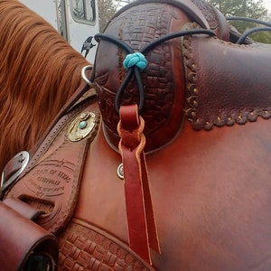 Op de afbeelding: Close-up van een bruin leren zadel met een turquoise en zilveren concho. Een zwarte band met een turquoise knoop is bevestigd aan de zadelboom.
