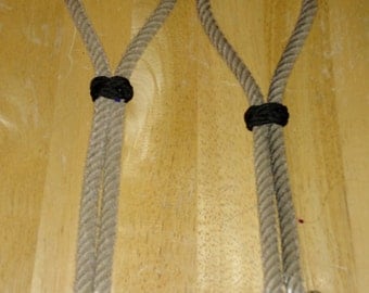Puede incluir: Dos restricciones de cuerda con anillos metálicos. Cada restricción presenta un lazo en la parte superior, un nudo negro y dos anillos metálicos en la parte inferior. Las cuerdas son de color beige claro.