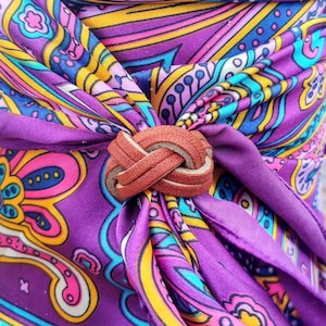 May include: A brown leather bolo tie with a knot design. The tie is tied around a purple and yellow paisley patterned scarf.