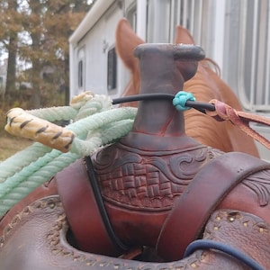 Op de afbeelding: Close-up van een bruin leren zadel met ingewikkelde houtsnijwerk. Een lichtblauw touw is om de zadelboom gewikkeld, en een bruin leren riem is aan de zadelboom bevestigd.