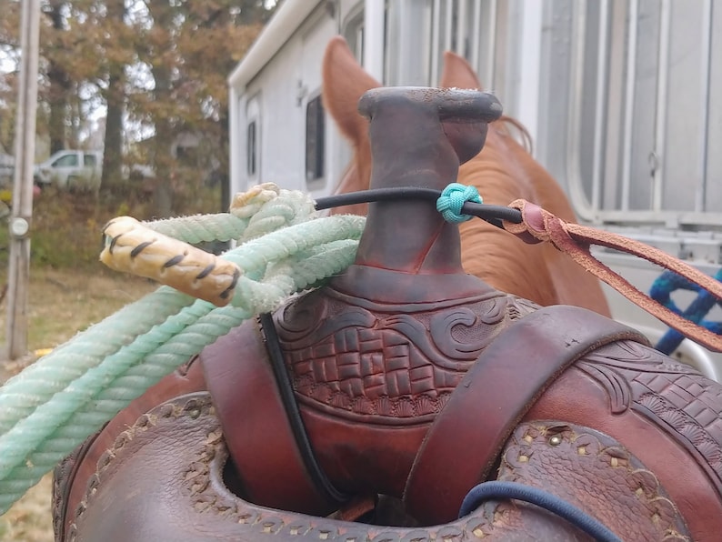 Op de afbeelding: Close-up van een bruin leren zadel met ingewikkelde houtsnijwerk. Een lichtblauw touw is om de zadelboom gewikkeld, en een bruin leren riem is aan de zadelboom bevestigd.