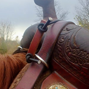 Op de afbeelding: Close-up van een bruin leren zadel met ingewikkelde houtsnijwerk en een turquoise accent. Een zwarte leren riem met een zilveren gesp is bevestigd aan het zadel.