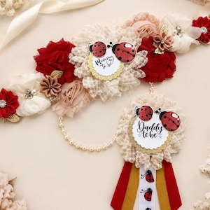 May include: A decorative sash and badge set featuring red and cream fabric flowers, gold leaves, and ladybug accents. The sash displays the text Mommy to be, while the hanging badge features the text Daddy to be and a series of ladybug icons.