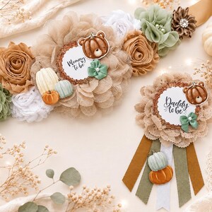 Pode incluir: Uma faixa de maternidade e um broche a condizer com flores de tecido bege, branco e verde sálvia. A faixa apresenta um pendente de abóbora em forma de urso e uma etiqueta com a inscrição Mommy to be. O broche tem uma abóbora urso e o texto Daddy to be.