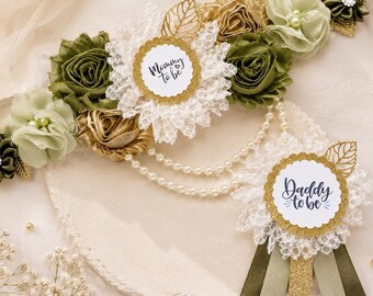 May include: A maternity sash featuring olive green and gold fabric flowers, white lace, and pearl strands, with a center badge reading Mommy to be. A matching pin badge features the text Daddy to be with olive green and gold ribbon streamers.