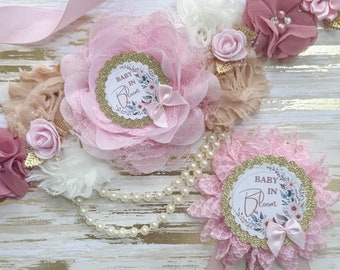 May include: Pink and gold baby shower sash with a large pink flower and a smaller pink flower with a gold "Baby in Bloom" sign. The sash is decorated with pearls and lace.