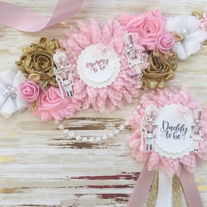 Puede incluir: Cintas de ducha nupcial rosa y dorada con temática de cascanueces. Las cintas presentan una roseta de encaje rosa y blanco con un círculo blanco que dice "Mommy to be" y una roseta de encaje rosa y blanco con un círculo blanco que dice "Daddy to be".