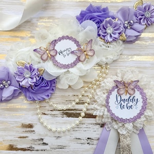 A beautifully designed baby shower accessory set in a lavender and ivory theme, featuring "Mommy to Be" and "Daddy to Be" badges. The set is decorated with delicate fabric flowers, lace, pearl strands, and gold accents. Glittery purple trim and elegant butterfly embellishments enhance the design, while cascading ribbon tails complete the sophisticated look. The accessories are displayed on a rustic wooden surface with gold detailing.