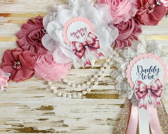 May include: Pink and white floral sash with a white lace trim and two fabric rosettes. The rosettes have a pink and white bow with the text "Mommy to be" and "Daddy to be" on them.