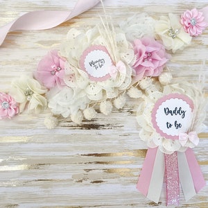 Op de afbeelding: Een roze en witte bloemenband met de tekst "Mommy to be" en een bijpassende roze en witte bloemenspeld met de tekst "Daddy to be".