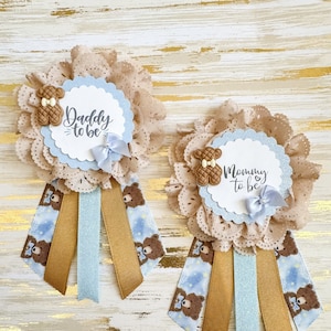 以下が含まれることがあります： テディベアのデザインが施された、茶色と青のベビーシャワーのサッシュが2つ。1つのサッシュには「Daddy to be」、もう1つのサッシュには「Mommy to be」と書かれています。