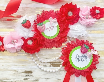 May include: Red and white satin sash with a strawberry and lace design. The sash has a green circle with the text "Mommy to be" and a pink bow. A matching sash with the text "Daddy to be" is also included.