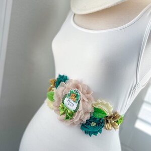 May include: A white maternity dress with a green, gold and blush floral sash. The sash features a  "Mommy to Be"  button with a cartoon lion.