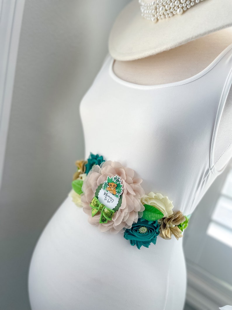 May include: A white maternity dress with a green, gold and blush floral sash. The sash features a  "Mommy to Be"  button with a cartoon lion.