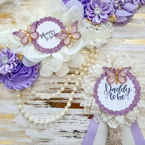 May include: Purple and white sash with a white lace overlay and a pearl necklace. The sash has a purple glitter circle with the text "Mommy to be" and two purple butterflies. The necklace has a purple glitter circle with the text "Daddy to be" and a purple butterfly.