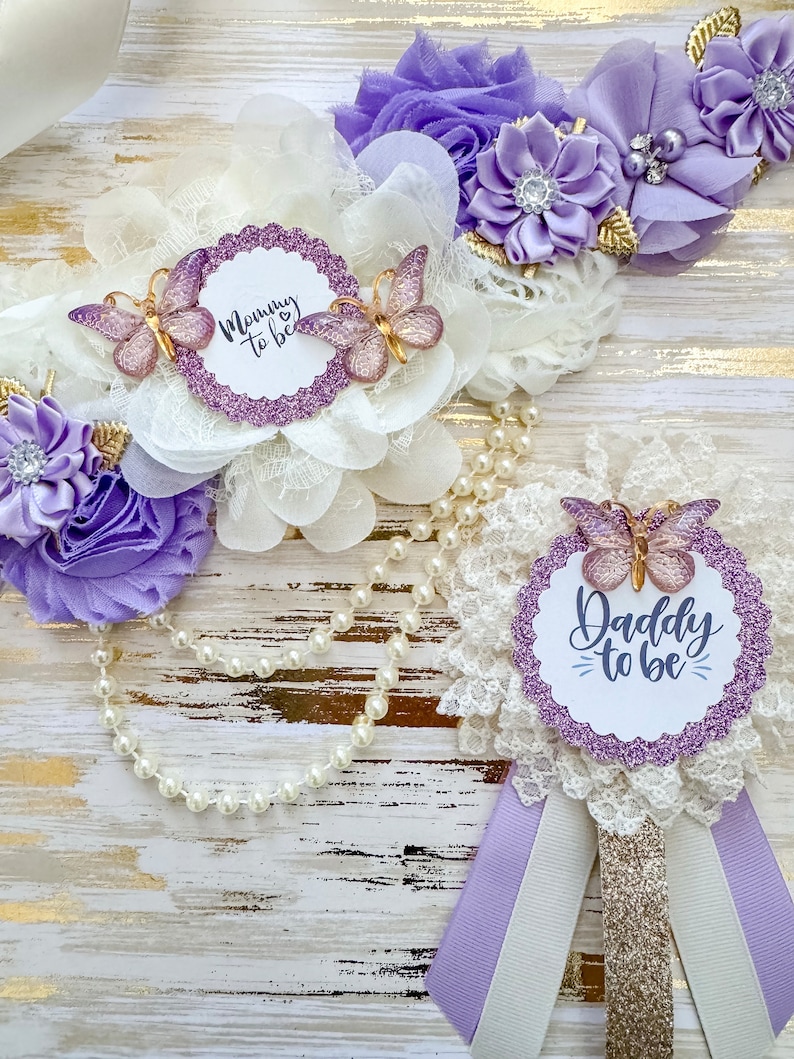 May include: Purple and white sash with a white lace overlay and a pearl necklace. The sash has a purple glitter circle with the text "Mommy to be" and two purple butterflies. The necklace has a purple glitter circle with the text "Daddy to be" and a purple butterfly.