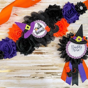Puede incluir: Una banda de baby shower con tem&aacute;tica de Halloween con flores negras, naranjas y moradas y un sombrero de bruja con el texto "A Baby is Brewing". Tambi&eacute;n se incluye un boutonniere a juego con un sombrero de bruja y el texto "Daddy to be".