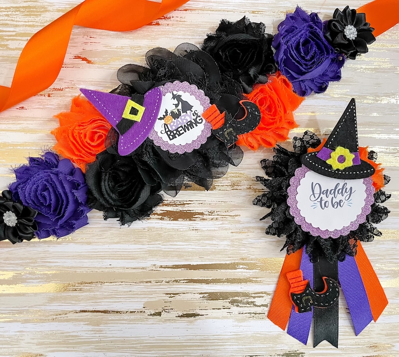 Puede incluir: Una banda de baby shower con tem&aacute;tica de Halloween con flores negras, naranjas y moradas y un sombrero de bruja con el texto "A Baby is Brewing". Tambi&eacute;n se incluye un boutonniere a juego con un sombrero de bruja y el texto "Daddy to be".
