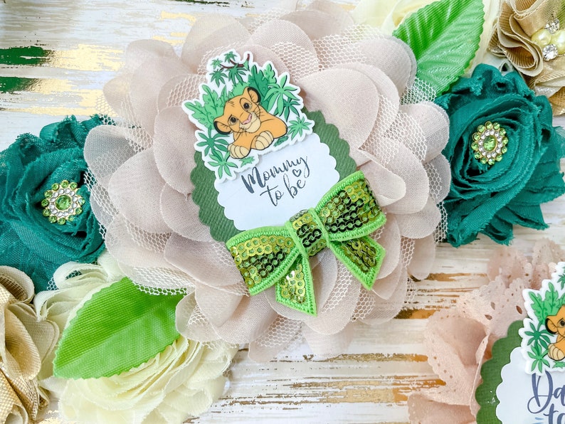 May include: A light brown and green floral headband with a green bow and a white circle with the text "Mommy to be" and a cartoon lion cub.