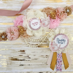 Puede incluir: Una faja floral rosa y dorada con una superposición de encaje blanco y dos rosetas de tela. Las rosetas tienen un diseño de taza de té y hada con el texto "Mommy to be" y "Daddy to be".