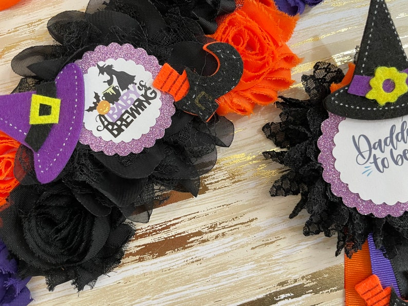 Puede incluir: Dos decoraciones para baby shower con tem&aacute;tica de Halloween. Una presenta un sombrero de bruja y el texto "A Baby is Brewing". La otra presenta un sombrero de bruja y el texto "Daddy to be".
