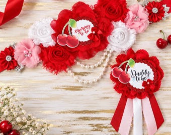 May include: A maternity sash and matching pin set featuring red, white, and pink fabric flowers, pearl strands, and cherry illustrations. The sash displays the text Mommy to be, while the pin features the text Daddy to be in script font.