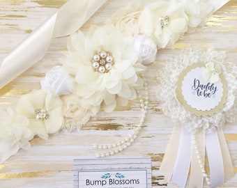 May include: A white satin sash with ivory and white flowers, pearls, and a rhinestone brooch. A white and ivory lace pin with a beige circle that says "Daddy to be" in blue script.