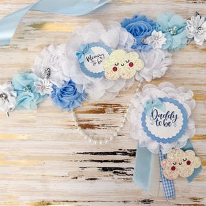 May include: A baby shower sash and pin set in white and blue. The sash features fabric flowers, a pearl strand, and a tag that says "Mommy to be". The pin says "Daddy to be" and has a cloud embellishment.
