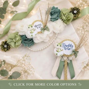 May include: A maternity sash and matching pin set featuring white and sage green fabric flowers, pearl strands, and gold-trimmed circular badges. The badges display a cartoon turtle hatching from an egg with the text Mommy to be and Daddy to be.
