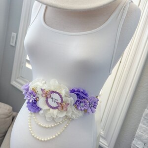 May include: A white maternity sash with purple and white flowers, a pearl strand, and a "Mommy to Be" sign with butterflies.