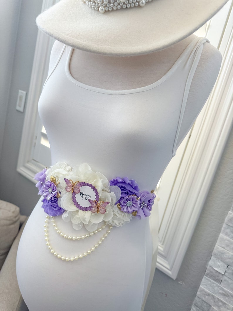 May include: A white maternity sash with purple and white flowers, a pearl strand, and a "Mommy to Be" sign with butterflies.