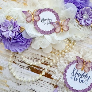 May include: Purple and white flower sashes with butterflies and glitter. The sashes say "Mommy to be" and "Daddy to be".