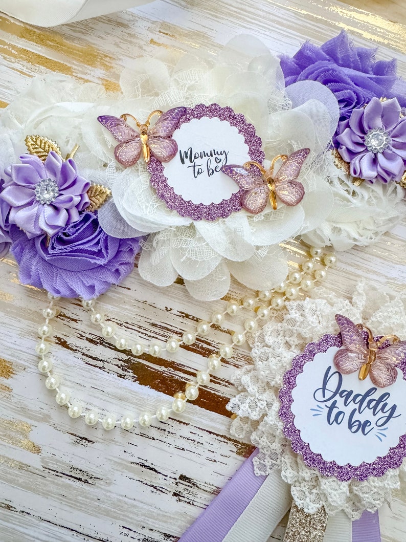 May include: Purple and white flower sashes with butterflies and glitter. The sashes say "Mommy to be" and "Daddy to be".