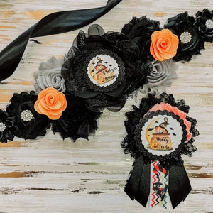 May include: A black and orange Halloween themed baby shower sash with the text "A Baby is Brewing" and a black and orange bow with the text "Daddy to Be".