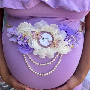 May include: Purple maternity sash with white flowers, a pearl necklace, and a sparkly pink circle with the text "Mommy to be".