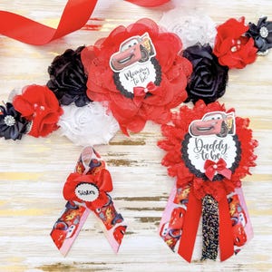 May include: A collection of baby shower decorations in red and black. The set includes a sash with the text "Mommy to be", a badge with "Daddy to be", and a ribbon with "Sister". Each item features a cartoon car design.
